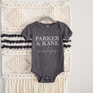 May include: A dark gray baby onesie with short sleeves and a snap closure. The onesie has the text "PARKER & KANE mockups" printed on the front in white.