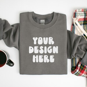May include: A gray sweatshirt with the text "YOUR DESIGN HERE" in white, paired with plaid pajama pants and a mug of coffee. The sweatshirt is folded, with the sleeves partially visible. The pajama pants have a red, green, and white plaid pattern.