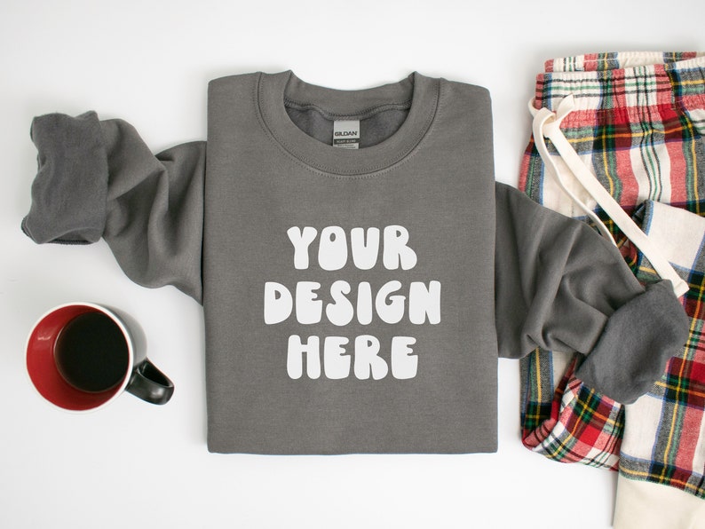 May include: A gray sweatshirt with the text "YOUR DESIGN HERE" in white, paired with plaid pajama pants and a mug of coffee. The sweatshirt is folded, with the sleeves partially visible. The pajama pants have a red, green, and white plaid pattern.
