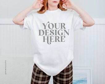 May include: White t-shirt with the text "YOUR DESIGN HERE" printed in gray on the front. The t-shirt is being worn by a person with long, red hair. The person is wearing brown and white plaid pants.