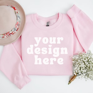 Puede incluir: Una sudadera rosa con el texto "your design here" en blanco. La sudadera está colocada sobre una superficie blanca con un sombrero marrón y un ramo de flores blancas.