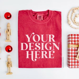 May include: A red t-shirt with the text "YOUR DESIGN HERE" printed in white. The t-shirt is surrounded by Christmas decorations, including nutcrackers, ornaments, and a red and white gingham gift box.