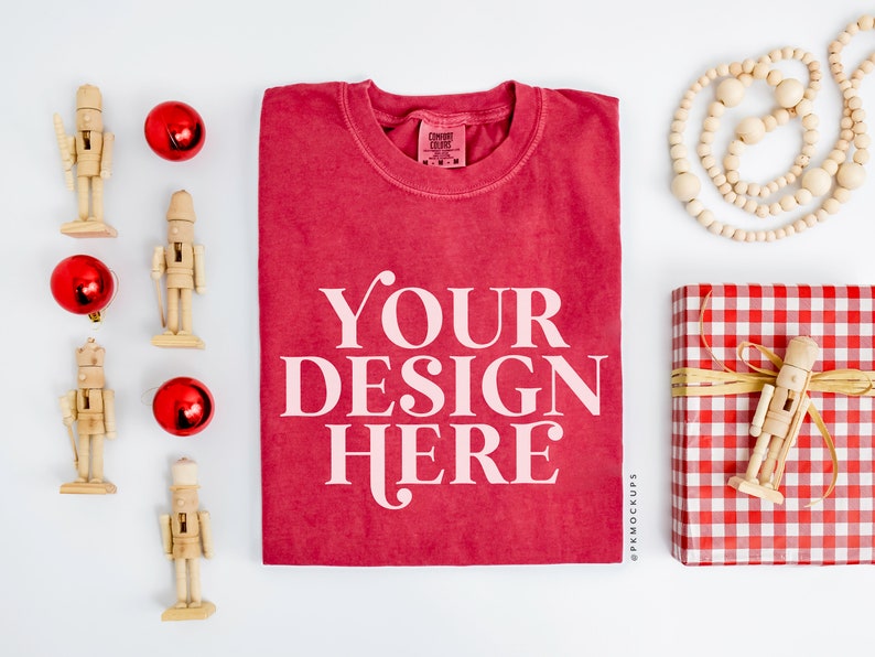 May include: A red t-shirt with the text "YOUR DESIGN HERE" printed in white. The t-shirt is surrounded by Christmas decorations, including nutcrackers, ornaments, and a red and white gingham gift box.