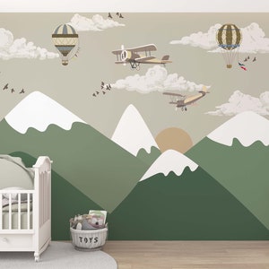 Op de afbeelding: Een muurschildering voor de kinderkamer met een berglandschap met groene en witte toppen, een ondergaande zon en cartoonvliegtuigen en luchtballonnen. Een witte wieg en een knuffelolifant staan op de voorgrond. Een mand met speelgoed staat op de grond.