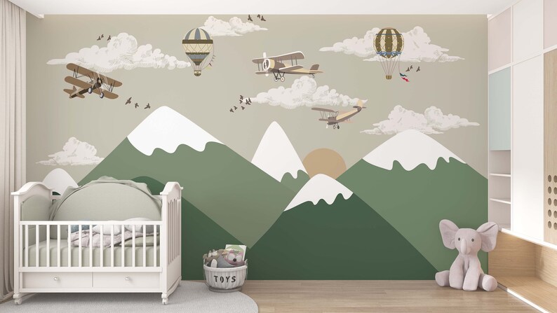 Op de afbeelding: Een muurschildering voor de kinderkamer met een berglandschap met groene en witte toppen, een ondergaande zon en cartoonvliegtuigen en luchtballonnen. Een witte wieg en een knuffelolifant staan op de voorgrond. Een mand met speelgoed staat op de grond.