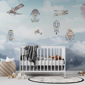 May include: A white crib with a gray pom-pom blanket, positioned against a wall mural featuring blue mountains, clouds, and vintage aircraft. A woven basket, wooden toys, and a round map-patterned rug are placed on the floor in front of the crib.
