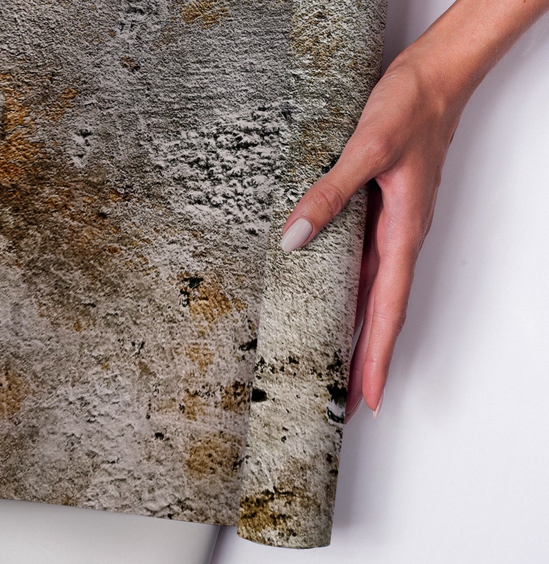 Puede incluir: Un rollo de papel pintado texturizado con apariencia de hormig&oacute;n envejecido. La paleta de colores incluye tonos de gris, marr&oacute;n y negro, creando una est&eacute;tica desgastada e industrial. El papel pintado est&aacute; siendo sostenido por una mano.