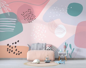 Puede incluir: Un mural de pared con formas abstractas en rosa, verde azulado y lavanda con patrones de líneas y puntos. Debajo, un cojín de suelo rosa claro, una almohada con forma de tiburón, una mesa auxiliar blanca con lámpara y barcos de juguete.
