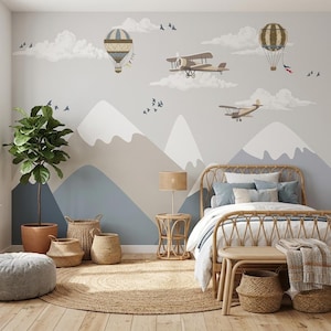 May include: A bedroom featuring a wall mural with mountain silhouettes, hot air balloons, and airplanes. The room includes a rattan bed frame with white bedding, a round jute rug, a small rattan side table with a lamp, and several woven baskets.