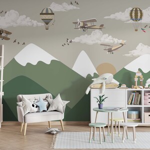 Op de afbeelding: Een kinderkamersc&egrave;ne met een muurschildering van bergen, wolken en vintage vliegtuigen en luchtballonnen. De kamer heeft een witte boekenkast, een kleine tafel en stoelen en een bank met stervormige kussens. Een walvisknuffel ligt op een kussen.