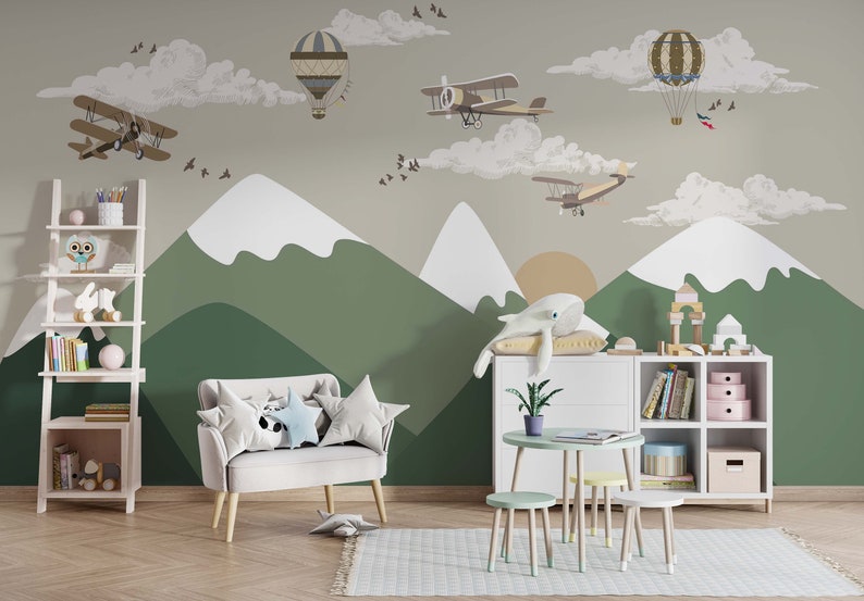 Op de afbeelding: Een kinderkamersc&egrave;ne met een muurschildering van bergen, wolken en vintage vliegtuigen en luchtballonnen. De kamer heeft een witte boekenkast, een kleine tafel en stoelen en een bank met stervormige kussens. Een walvisknuffel ligt op een kussen.