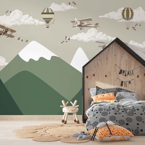 Op de afbeelding: Een kinderkamer met een muurschildering van bergen, wolken, vliegtuigen en luchtballonnen. Een houten bedframe in de vorm van een huis met de tekst "DREAM ON" staat op de voorgrond, samen met een kleine stoel en een rond tapijt.