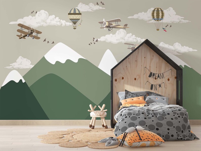 Op de afbeelding: Een kinderkamer met een muurschildering van bergen, wolken, vliegtuigen en luchtballonnen. Een houten bedframe in de vorm van een huis met de tekst "DREAM ON" staat op de voorgrond, samen met een kleine stoel en een rond tapijt.