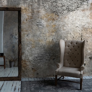 Puede incluir: Un sill&oacute;n alto tapizado en beige con respaldo abotonado se encuentra contra una pared texturizada. Un gran espejo con marco de madera refleja una habitaci&oacute;n con un peque&ntilde;o taburete y un jarr&oacute;n. Una alfombra estampada descansa sobre el suelo de madera.