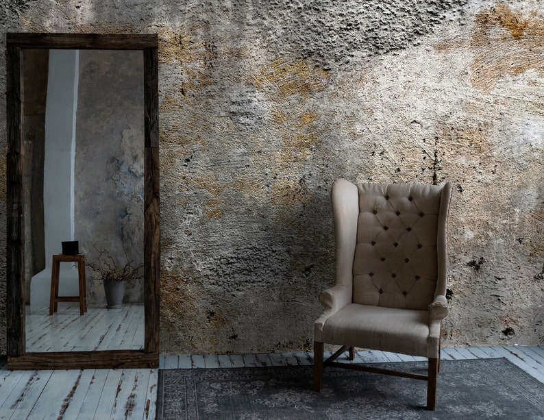Puede incluir: Un sill&oacute;n alto tapizado en beige con respaldo abotonado se encuentra contra una pared texturizada. Un gran espejo con marco de madera refleja una habitaci&oacute;n con un peque&ntilde;o taburete y un jarr&oacute;n. Una alfombra estampada descansa sobre el suelo de madera.