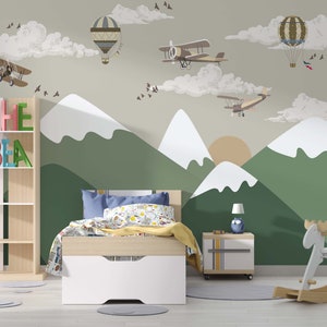 Op de afbeelding: Kinderslaapkamer met een berg- en luchtmuurschildering met vliegtuigen en luchtballonnen. De kamer heeft een bed, een nachtkastje, een boekenplank met de naam "CHE BIA" en een houten schommelende eland.