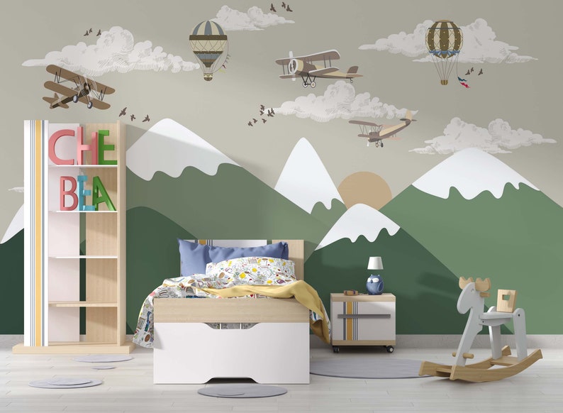 Op de afbeelding: Kinderslaapkamer met een berg- en luchtmuurschildering met vliegtuigen en luchtballonnen. De kamer heeft een bed, een nachtkastje, een boekenplank met de naam "CHE BIA" en een houten schommelende eland.