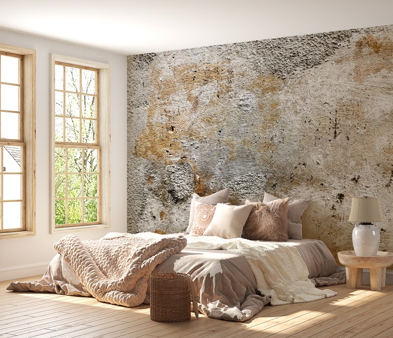 Puede incluir: Dormitorio con una cama en tonos neutros. La cama tiene almohadas, una manta de punto beige y una cesta de mimbre. Grandes ventanas y una pared texturizada completan la escena, creando un ambiente tranquilo.