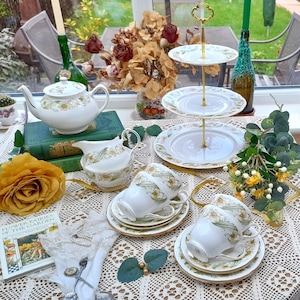 May include: A three-tiered cake stand with white porcelain plates and gold trim. The stand is decorated with floral patterns and is filled with teacups and a teapot. The stand is on a white tablecloth with a yellow rose and a book titled "Flower Fairies of the Garden".
