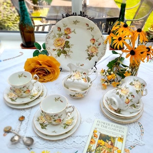 May include: A vintage tea set with a floral pattern featuring yellow roses. The set includes a teapot, creamer, sugar bowl, and two teacups and saucers. The tea set is displayed on a white tablecloth with a lace pattern. A book titled "Flower Fairies of the Garden" by Cicely Mary Barker is also visible.