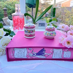 May include: A pink decorative book with the title "The Pink Fairy Book" and two small, ornate, lidded jars with floral designs. The jars sit on top of the book, surrounded by white roses and pink flowers.