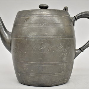 May include: Antique pewter teapot with a barrel-shaped body and a curved spout. The teapot features a handle, a lid with a small knob, and horizontal bands around the body. The text "Rose Club R.P." is inscribed on the side.