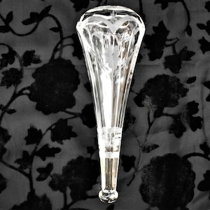 May include: A clear, etched glass bottle stopper with a flared top and a slender, elongated body. The stopper features a floral design and is set against a black background with a floral pattern.