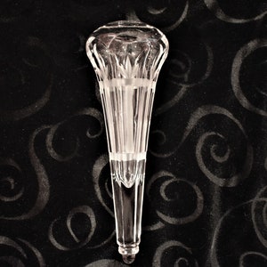 May include: A clear, cut-glass vase with a flared top and a long, slender stem. The vase features vertical cuts and etched floral designs. The vase is displayed against a black background with a swirling pattern.
