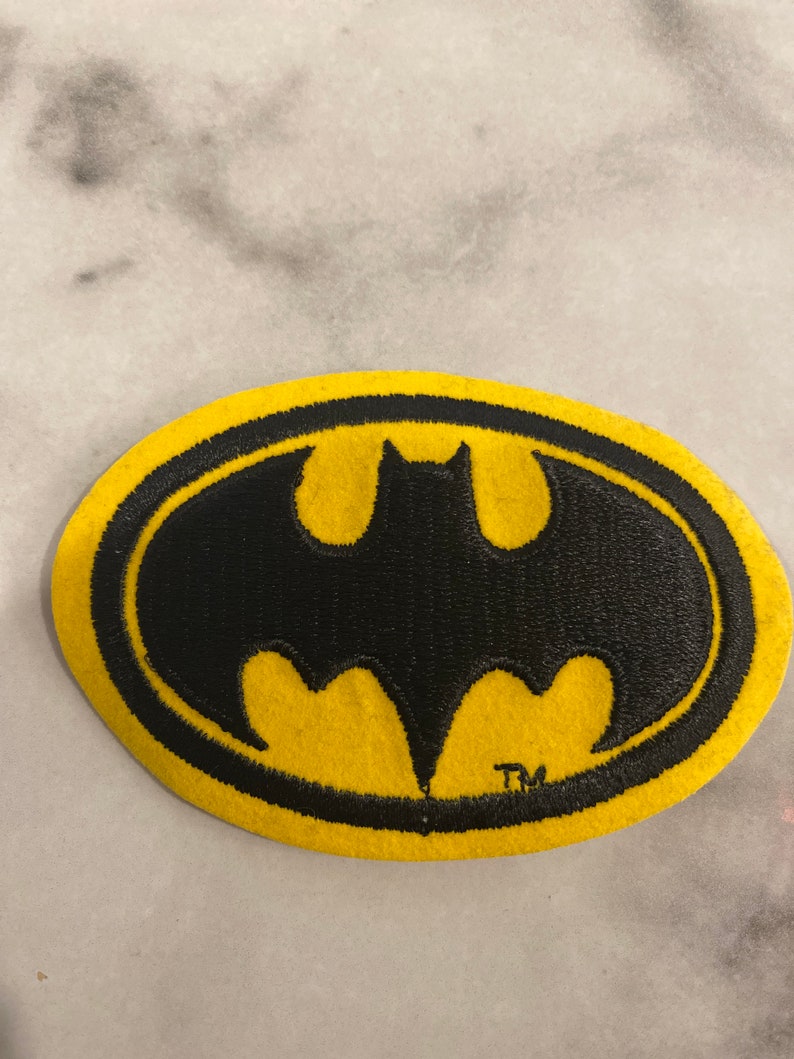 Op de afbeelding: Een geel en zwart Batman logo-patch. De patch toont het iconische vleermuissymbool in zwart, tegen een gele ovale achtergrond. Het handelsmerksymbool is rechtsonder zichtbaar.