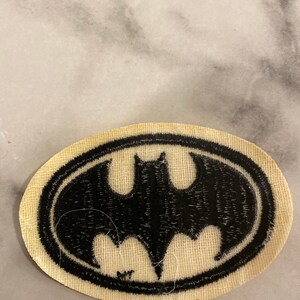 Op de afbeelding: Een ovaalvormige patch met het Batman-logo. Het zwarte vleermuissymbool is omlijnd door een zwarte rand, tegen een beige stoffen achtergrond. Deze patch is ontworpen voor toepassing op kleding of accessoires.