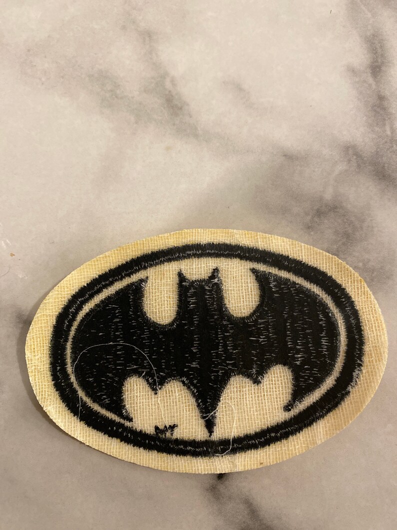 Op de afbeelding: Een ovaalvormige patch met het Batman-logo. Het zwarte vleermuissymbool is omlijnd door een zwarte rand, tegen een beige stoffen achtergrond. Deze patch is ontworpen voor toepassing op kleding of accessoires.
