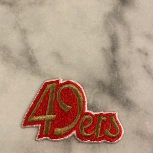 Può includere: Toppa ricamata con la scritta "49ers" in oro e rosso. La toppa ha un bordo bianco ed è probabilmente per una squadra sportiva. La toppa è su una superficie di marmo.