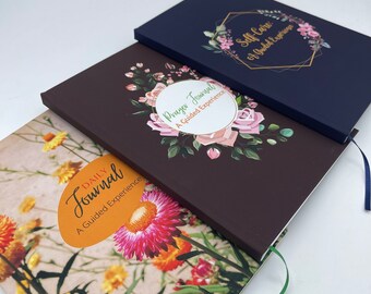 May include: Three journals with floral designs and gold accents. The top journal is navy blue with the text "Self-Care: A Guided Experience". The middle journal is brown with the text "Prayer Journal: A Guided Experience". The bottom journal is yellow with the text "Daily Journal: A Guided Experience".