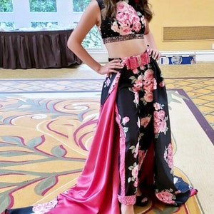 May include: A woman wearing a black and pink floral print two-piece outfit with a pink satin train. The outfit features a crop top and pants with a matching belt.