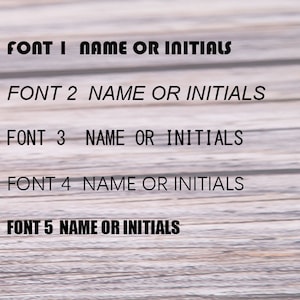 May include: Image showcasing five different font styles labeled Font 1 through Font 5. Each font is accompanied by the text "NAME OR INITIALS" in black, against a light wood grain background. The fonts vary in style, offering a selection for personalization.