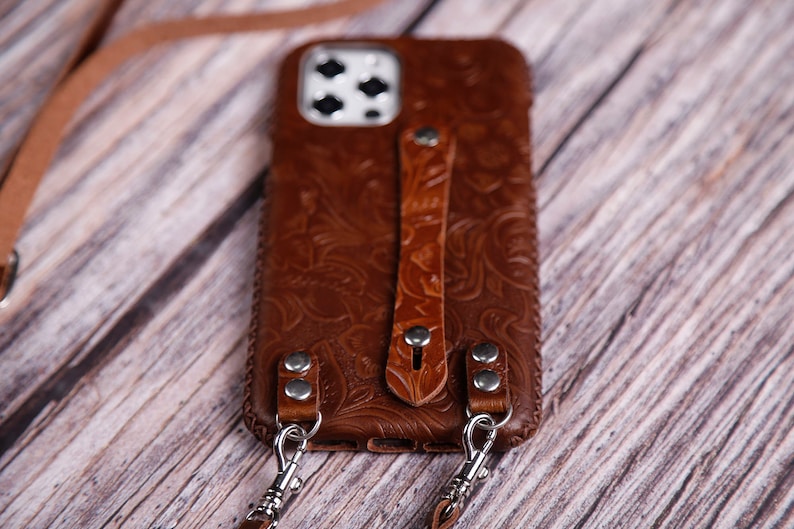 May include: A brown leather phone case with a floral embossed design. The case has a matching leather strap and silver-tone metal hardware. The case is designed to hold a smartphone and has a strap for carrying.