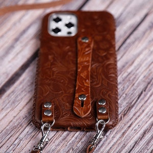 May include: A brown leather phone case with a floral pattern and a strap. The case has two metal clasps for attaching a strap or chain.
