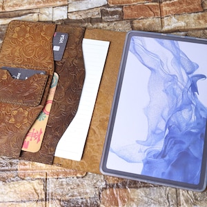 May include: A brown leather folio case with a floral embossed design. The case has a slot for a credit card and a pocket for a pen. The case is open to reveal a white notepad and a tablet with a blue abstract design.