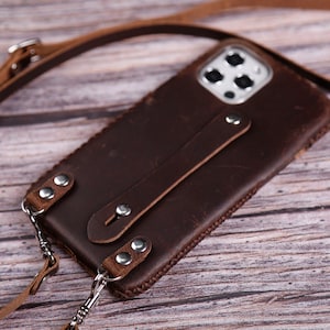 May include: A brown leather phone case with a matching strap. The case has a stitched border and silver-tone rivets. The strap is attached with silver-tone clasps and is adjustable. The phone case is designed to fit a smartphone.