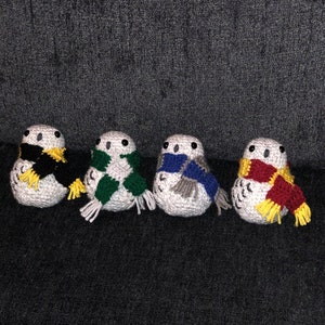 May include: Four handmade crochet owls with white bodies and black eyes. Each owl wears a scarf in different colors: black and yellow, green and white, blue and gray, and red and yellow. The owls are arranged in a row on a dark gray surface.