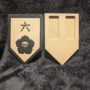 May include: A tan-colored wooden plaque with a black border and a black Japanese character and flower design. The back of the plaque is also tan-colored and has two rectangular cutouts.
