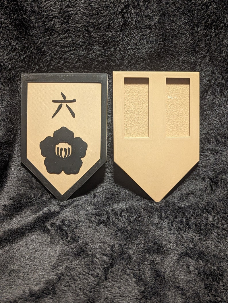 May include: A tan-colored wooden plaque with a black border and a black Japanese character and flower design. The back of the plaque is also tan-colored and has two rectangular cutouts.