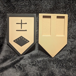 May include: A beige and black wooden shield with a black border and a black symbol in the center. The symbol is a black square with a black cross and a black diamond with white stars inside. The shield is attached to a beige wooden stand with two slots.