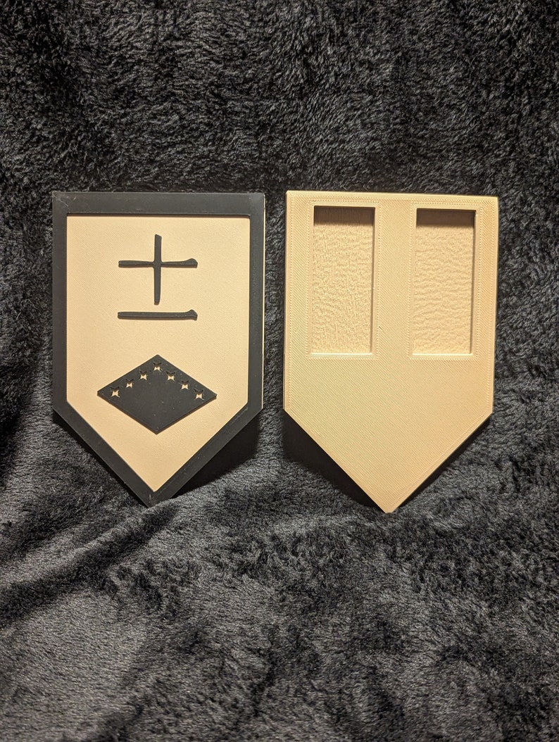 May include: A beige and black wooden shield with a black border and a black symbol in the center. The symbol is a black square with a black cross and a black diamond with white stars inside. The shield is attached to a beige wooden stand with two slots.