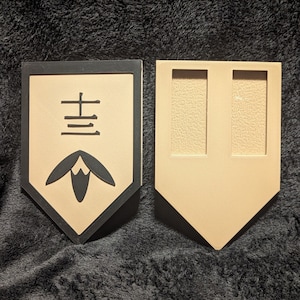 May include: A black and tan wooden plaque with a black border and a Japanese character in the center. The character is surrounded by a stylized flower design. The plaque is a decorative item.