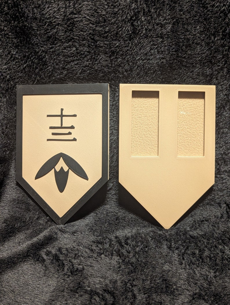 May include: A black and tan wooden plaque with a black border and a Japanese character in the center. The character is surrounded by a stylized flower design. The plaque is a decorative item.