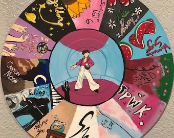 May include: A vinyl record painted with a colorful design featuring a person in a pink shirt and blue jeans, a sunflower, a cherry, a watermelon, and the words "Sunflower Vol. 6", "Cherry", "Watermelon Sugar", "To Be", "PWK", "Falling", "Canyon Moon", "Lights Up", "Golden", "Adore You", "Fine Line", and "She".