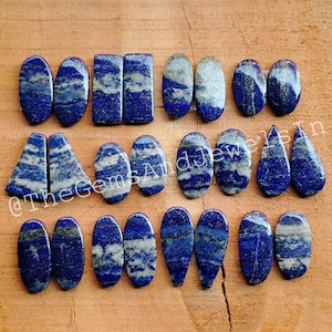 Könnte beinhalten: Eine Sammlung polierter Lapislazuli-Edelsteine in verschiedenen Formen, darunter Ovale, Rechtecke und Tropfen. Die Steine zeigen eine tiefblaue Farbe mit weißer und grauer Marmorierung, auf einer Holzoberfläche.