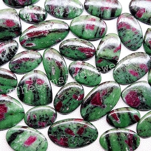May include: A collection of polished ruby zoisite gemstones. The stones are oval and oblong, with a mix of green and red colors. The gemstones are smooth and have a glossy finish. The text "theGems7ndustry" is visible on some of the stones.