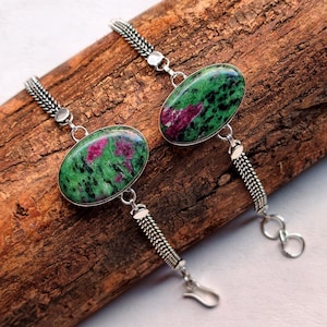 Op de afbeelding: Twee zilveren armbanden met ovale edelstenen. De edelstenen zijn groen met rode insluitsels. De armbanden hebben een kettingontwerp en een haaksluiting. Ze liggen op een houten oppervlak.